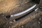 Curved steel cavalry sabre with rune line, ribbed black grip and its black scabbard on a weathered stone step