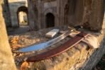 Curved steel scimitar on a sunlit stone ledge with a tooled leather scabbard, sharpening stone and cloth