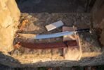 Curved steel scimitar seen from above on a stone sill beside its tooled brown leather scabbard and a whetstone