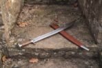 Curved steel tulwar with wood grip and disc pommel lying across its russet leather scabbard on stone steps
