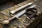 Damascus arming sword resting on a lichen covered brick wall with the coiled brown leather belt and rune stamped scabbard beside it