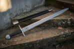 Damascus bastard sword with etched Valknut panel and long black grip beside its black scabbard on old stone steps