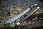 Damascus falchion sword with brass disc guard and dark wood grip beside its wooden scabbard on an old brick wall
