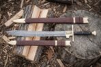 Damascus falchion with brass cross guard, black grip and brass pommel laid on a log between two brown leather scabbard pieces