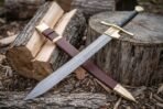 Damascus falchion with brass cross guard crossed over its brown leather scabbard with brass chape on split firewood logs