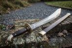 Damascus falchion with brass disc guard, hooked brass pommel and dark wood grip next to its wooden scabbard on brick