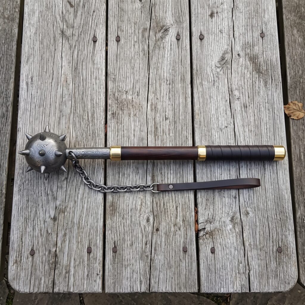 Damascus flail with a forge welded spiked ball head, brass collars, rosewood haft and hanging chain laid on a silvered timber table top
