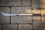 Damascus glaive with a ladder patterned forge welded blade and pale ash haft with a leather wrapped grip lying on grey cobbled flagstones