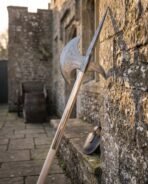Damascus halberd head with crescent axe blade and spike on a pale ash haft leaning against a sunlit castle wall