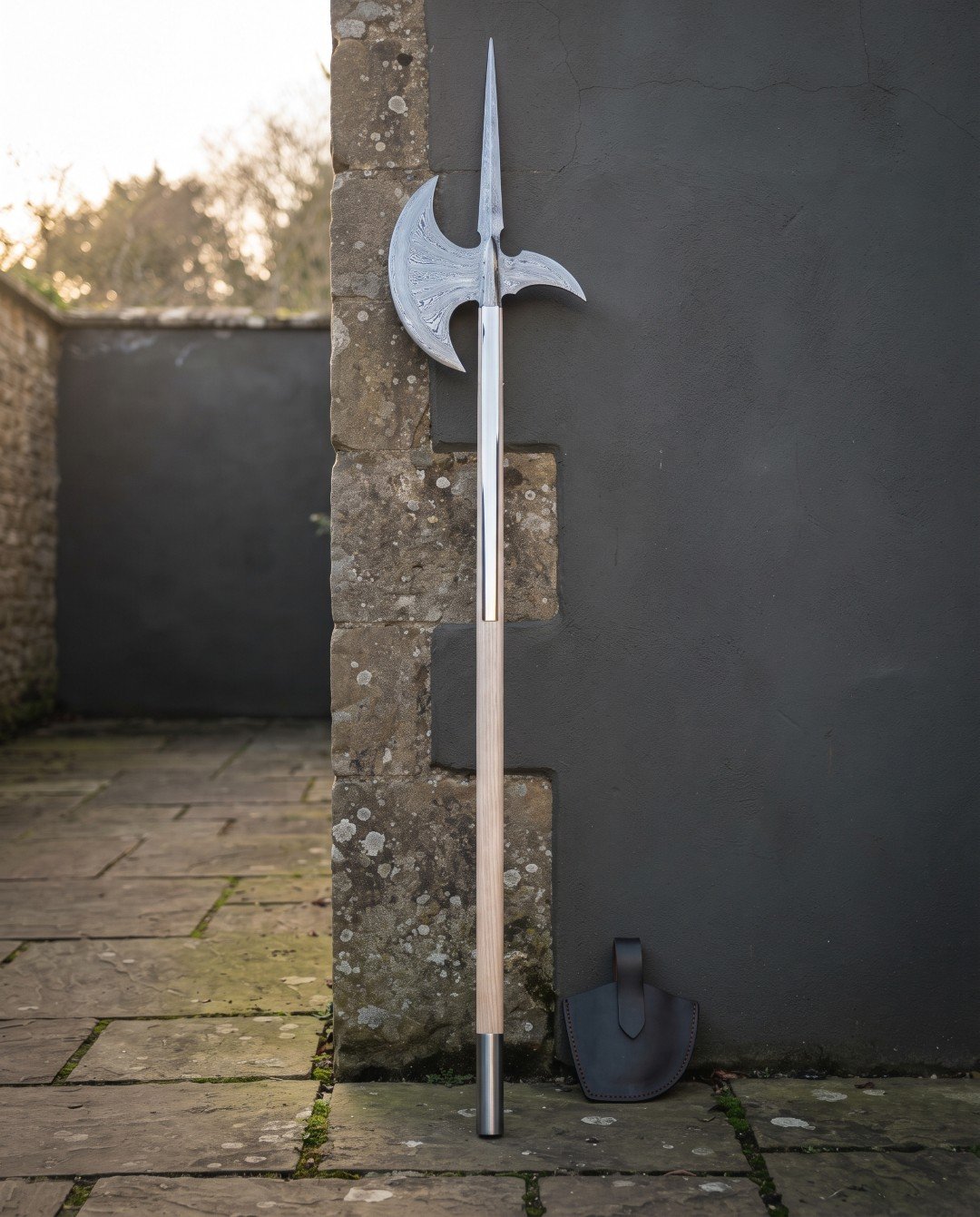Damascus halberd with a forge welded spike, patterned crescent blade and hooked fluke on a pale ash haft standing agains Damascus halberd with a forge welded spike, patterned crescent blade and hooked fluke on a pale ash haft standing against a stone and slate garden wall