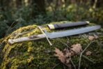 Damascus langes messer with stag horn grip, brass cross guard and pommel on a mossy rock beside its black leather scabbard