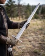 Damascus longsword with steel cross and Valknut pommel held flat across gloved hands in a dry meadow
