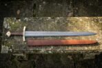 Damascus medieval sword with brass cross, faceted brass pommel and wooden grip on a mossy stone slab beside its leather scabbard