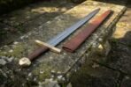 Damascus medieval sword with brass hilt and brown wooden grip next to its tan leather scabbard on a weathered stone bench