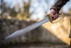 Damascus naval cutlass with brass shell guard held blade out against a blurred castle wall in low winter sun