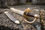 Damascus naval cutlass with brass shell guard, rune engraved knuckle bow and dark ribbed grip beside its scabbard on stone