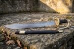 Damascus naval cutlass with rune etched blade and brass hilt lying above its black scabbard on a stone ledge