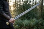 Damascus Norse sword with black grip and brass fittings held out at arm length in a sunlit forest