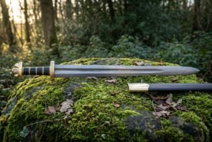Damascus Norse sword with dark etched layered steel blade and leather wrapped grip resting on a moss covered boulder in green woodland light