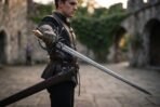 Damascus rapier with a swept hilt held out at full reach by a young man in a stone castle courtyard, scabbard at his hip