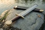 Damascus short sword with leather wrapped grip and brass Vegvisir pommel crossed over its scabbard on a lakeside rock