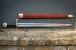 Damascus steel arming sword with straight brass cross and disc pommel laid on a stone ledge below its red leather scabbard