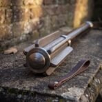 Damascus steel bar mace with a dark steel haft resting on weathered stone beside a looped brown leather strap and brick wall