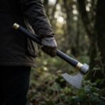 Damascus steel bearded axe with a black leather wrapped haft and brass cap, held blade down in a gloved hand in woodland