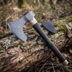 Close up of a Damascus steel bearded axe head with a brass collar, resting against a mossy log with a black leather blade sheath