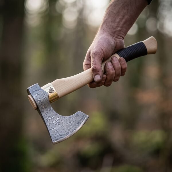 Damascus steel bearded axe with a pale wood haft and dark leather grip wrap held in one hand in a misty forest