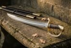Damascus steel cavalry sabre with brass hilt and ribbed grip lying on a mossy stone ledge beside its black scabbard
