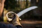 Damascus steel cavalry sabre with brass knuckle guard and ribbed grip held aloft by a gloved hand in dim woodland