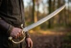 Damascus steel curved sabre with brass basket guard held low at the hip, blade angled up against a forest background