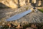Damascus steel cutlass seen point first along a mossy stone wall, showing the layered pattern on its wide blade