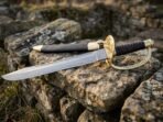 Damascus steel cutlass with brass shell guard, rune engraved knuckle bow and dark leather grip laid across lichen covered stones