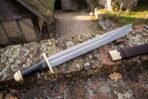 Damascus steel falchion with rune engraved blade, brass guard and pommel and black grip resting on a lichen covered brick wall