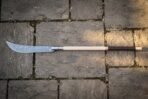 Damascus steel glaive with a curved single edged blade on a pale ash haft laid across sunlit flagstones