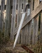 Damascus steel longsword leaning on a grey picket fence with its brown leather scabbard lying in the grass below