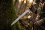 Damascus steel messer with brass crossguard and dark wooden grip held blade out in front of a mossy tree