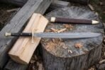 Damascus steel messer with brass crossguard and pommel cap laid flat on a chopping stump with its brown sheath behind