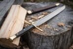 Damascus steel messer with brass fittings and black grip resting on a split log and stump beside its leather sheath