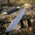 Damascus steel pirate cutlass with broad patterned blade and brass shell guard resting on a dry stone wall beside its scabbard