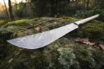 Close up of a Damascus steel polearm blade with a curved edge and steel collar, resting on a moss covered rock in autumn woodland