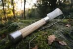 Damascus steel polearm laid on a mossy rock, pale wooden haft with a polished steel butt cap in the foreground and the blade behind