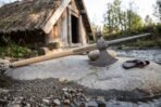 Damascus steel poleaxe head with spike and hammer poll on a pale ash haft resting on a river boulder by a thatched hut