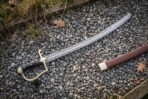 Damascus steel Polish szabla with open brass hilt and black horn grip laid on grey gravel beside its brown leather scabbard
