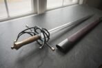 Damascus steel rapier with a blackened swept hilt and brass fittings on a slate windowsill beside its dark leather scabbard