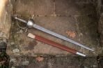 Damascus steel rapier with polished cup hilt and wire wrapped grip lying on flagstones beside a tan leather scabbard