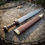 Damascus steel Roman gladius laid on a weathered tree stump next to its brown leather scabbard with brass throat and chape