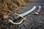 Damascus steel sabre with brass rune hilt laid on a gravel path with its brass tipped leather scabbard in the background